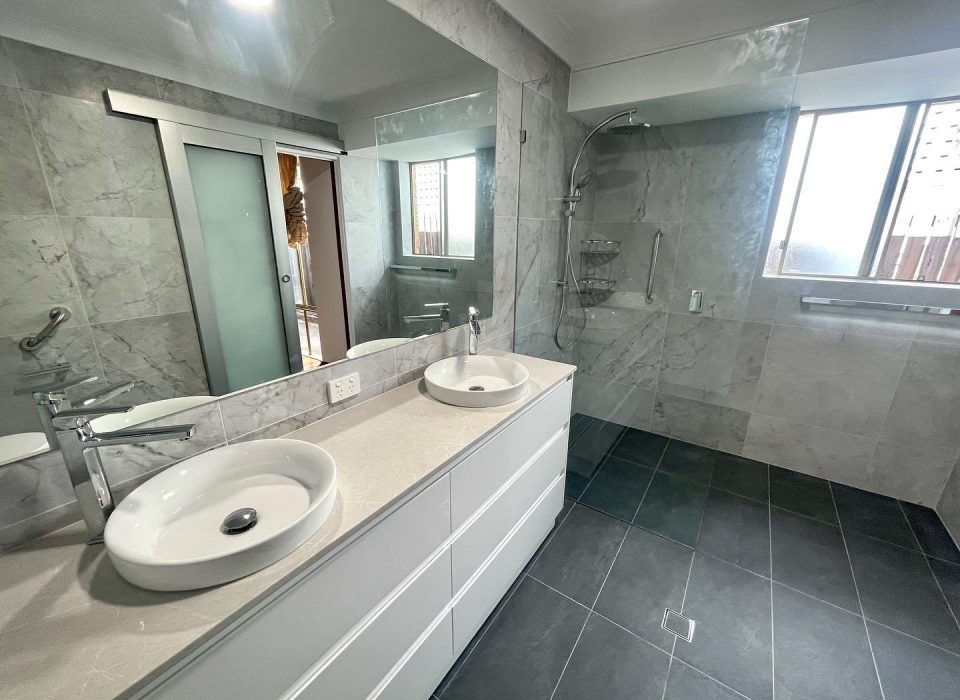 A renovated ensuite bathroom featuring large matte grey floor tiling, white marble wall tiles, a large mirror, double vanity and walk-in shower
