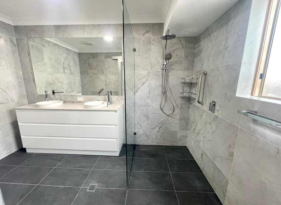 A renovated ensuite bathroom featuring large matte grey floor tiling, white marble wall tiles, a large mirror, double vanity and walk-in shower