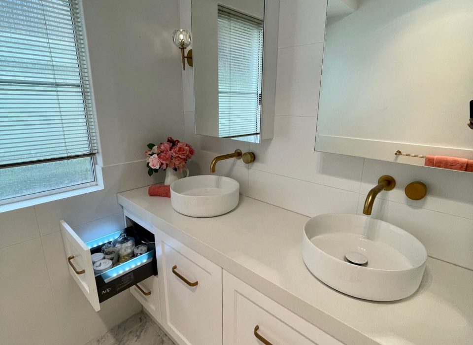 A renovated bathroom with white marble flooring, a double vanity with white cabinetry, basin sinks, mirrored medicine cabinets, and brass tapware