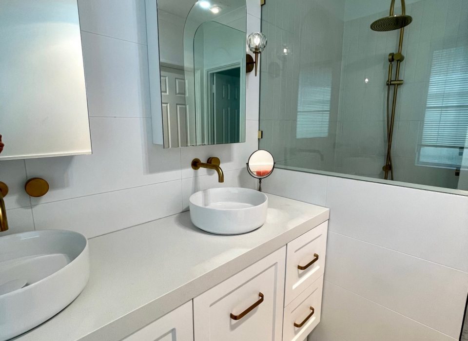 A renovated bathroom with white marble flooring, a double vanity with white cabinetry, basin sinks, mirrored medicine cabinets, brass tapware, and a shower enclosure