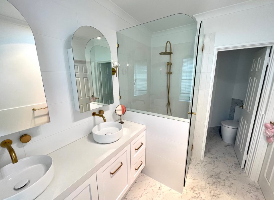 A renovated bathroom with white marble flooring, a double vanity with white cabinetry, basin sinks, mirrored medicine cabinets, brass tapware, and a shower enclosure