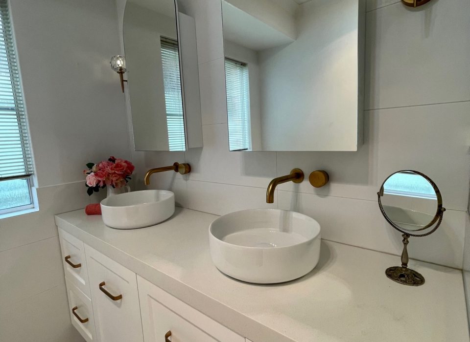 A renovated bathroom with white marble flooring, a double vanity with white cabinetry, basin sinks, mirrored medicine cabinets, and brass tapware