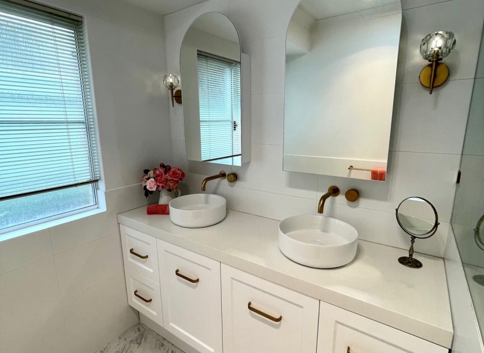 A renovated bathroom with white marble flooring, a double vanity with white cabinetry, basin sinks, mirrored medicine cabinets, and brass tapware