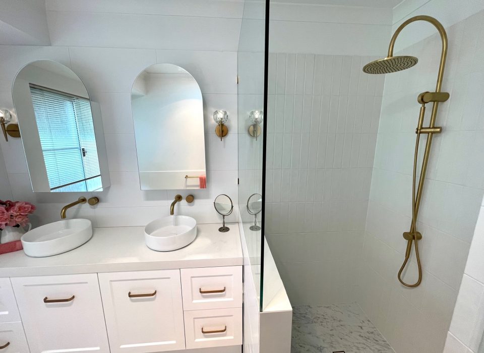 A renovated bathroom with white marble flooring, a double vanity with white cabinetry, basin sinks, mirrored medicine cabinets, brass tapware, and a shower enclosure