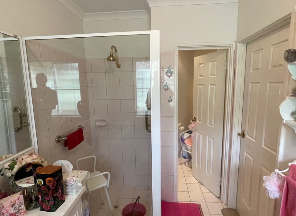 A cluttered bathroom with cream floor tiling, a pink bathmat and towels, shower enclosure, and plain white vanity