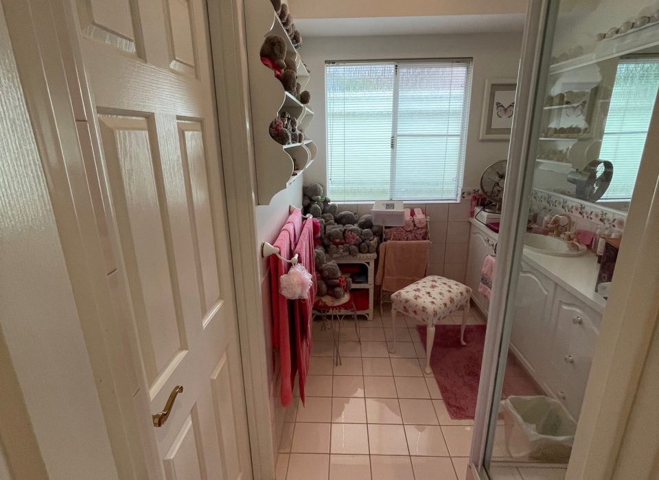 A cluttered bathroom with cream floor tiling, a pink bathmat and towels, shower enclosure, and plain white vanity