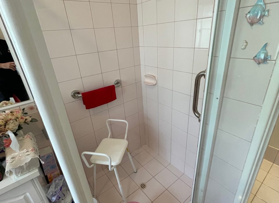 A cluttered bathroom with cream floor tiling, a pink bathmat, and shower enclosure with a gold shower head