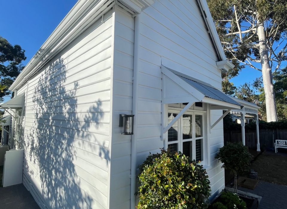 A worker's cottage-style home that is painted all white and framed with dwarf trees