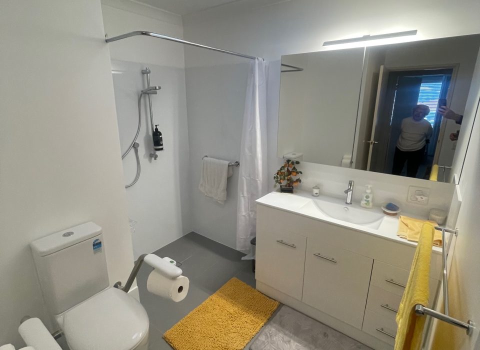 A renovated bathroom with an accessible toilet suite, shower, sink vanity, yellow bathmat and towel