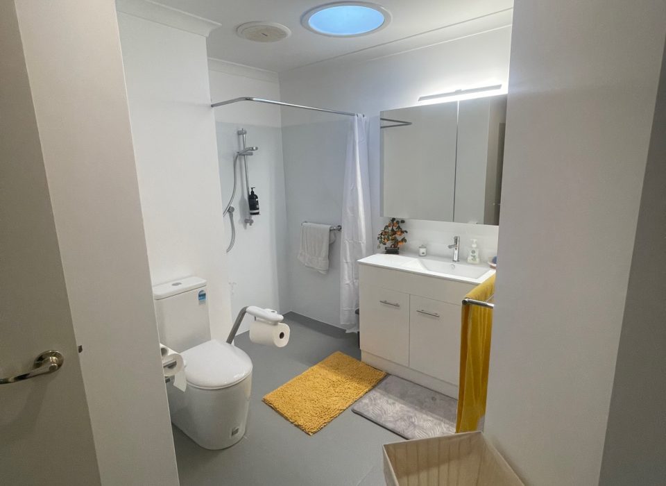 A renovated bathroom with an accessible toilet suite, shower, sink vanity, yellow bathmat and towel