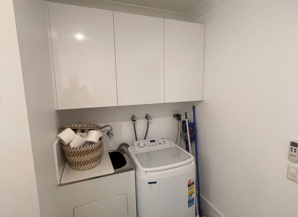 A renovated laundry with a sink, three overhead cupboards, a washing machine, and cleaning equipment stacked in the corner