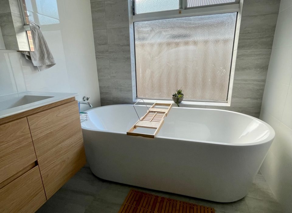 A renovated bathroom with a floating wood-look vanity, freestanding bathtub, and window