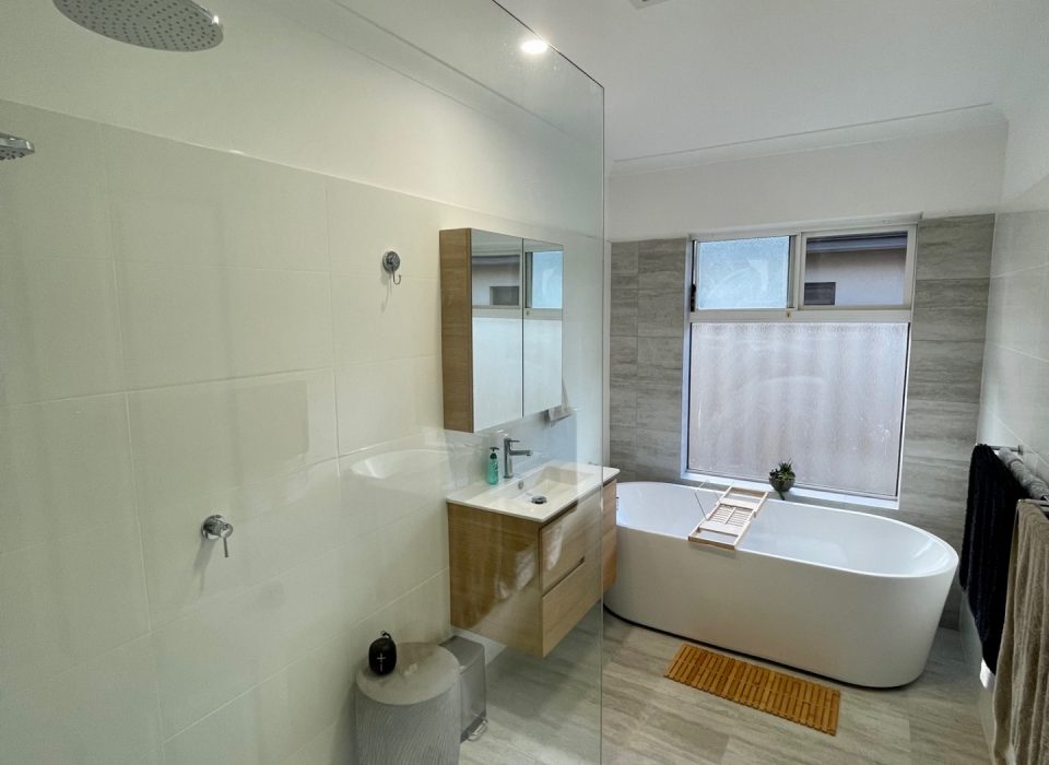 A renovated bathroom with a floating wood-look vanity, freestanding bathtub, and window