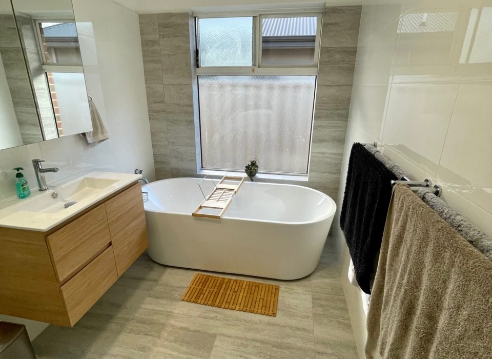A renovated bathroom with a floating wood-look vanity, freestanding bathtub, and window