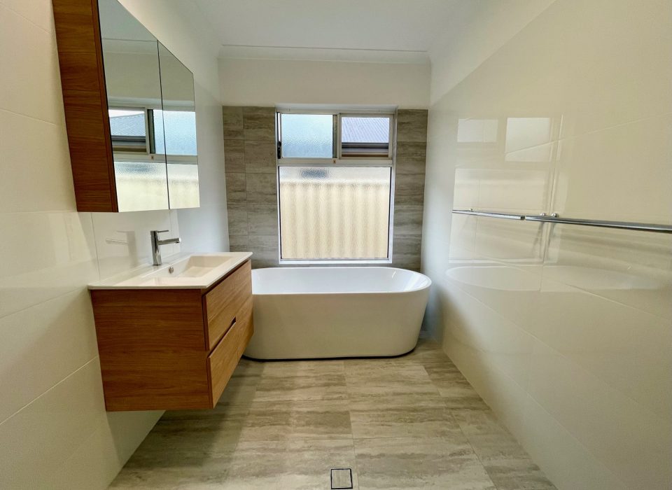 A renovated bathroom with a floating wood-look vanity, freestanding bathtub, and window