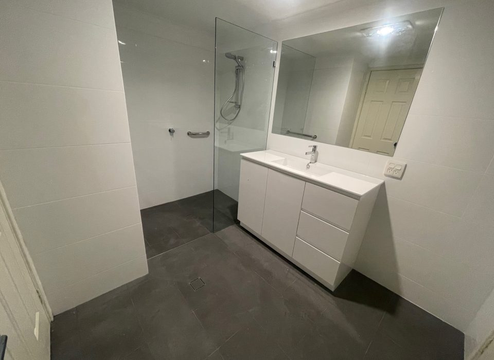 A modern renovated bathroom with dark gray matte floor tiling, an all-white vanity, walk-in shower, and large mirror