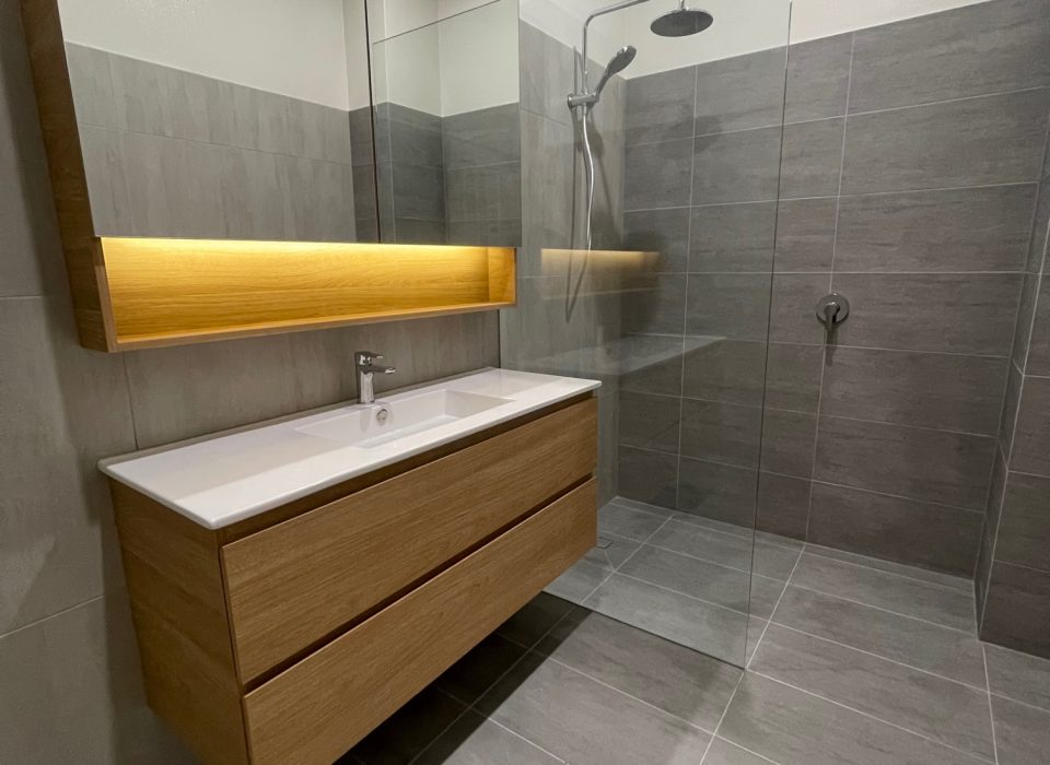 A modern bathroom with a walk-in shower, floating vanity with a large mirrored cabinet, chrome tapware, and large grey floor and wall tiling