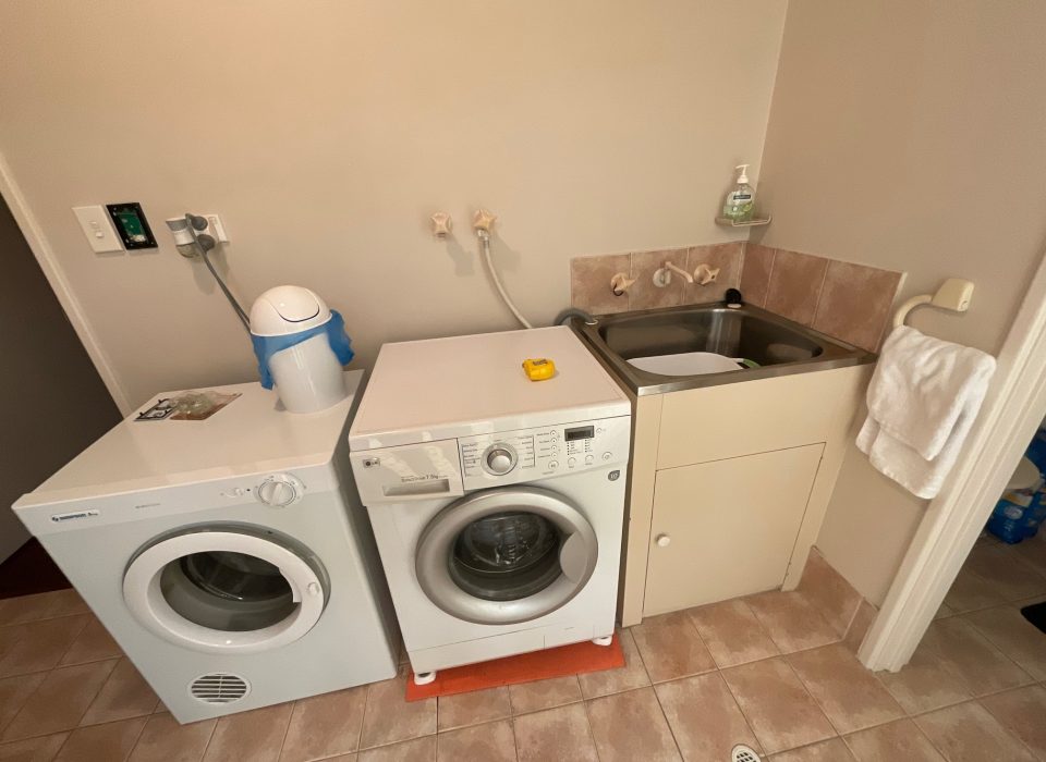 An old laundry space which features a washer and dryer with exposed plugs and hoses, next to a worn laundry sink